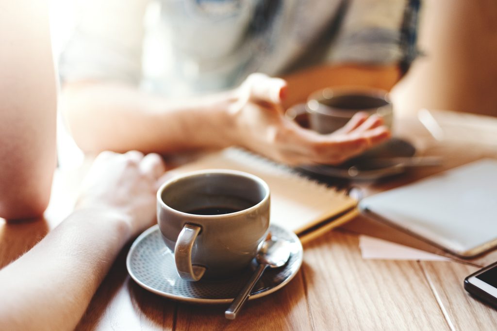 Friends talking at cafe table during coffee break. Unrecognizable male and female colleagues discussing business issues, focus on coffee cup with saucer and teaspoon | Pelican Coffee House